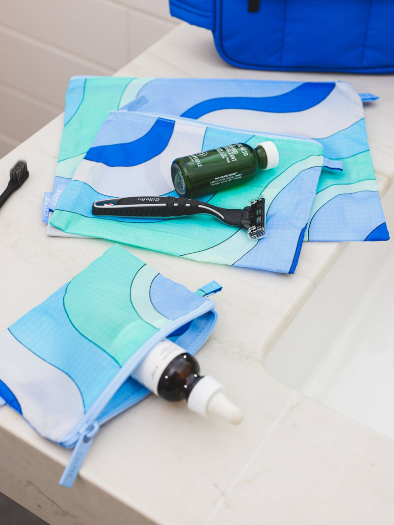 From CALPAK's Compakt collection, these 'Groovy Blue' zippered nylon pouches are displayed on a bathroom sink, offering stylish organization for your essentials