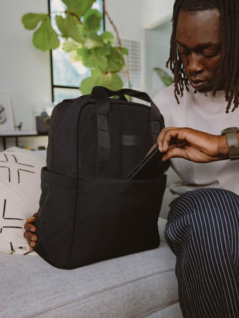 Model placing cell phone in front pocket of CALPAK Connect Laptop Backpack in black