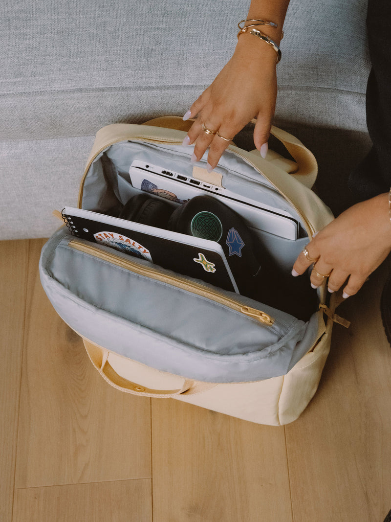 Model shows displays interior of sunshine CALPAK Connect Laptop Backpack with laptop in padded laptop sleeve and headphones and notebook in the main compartment