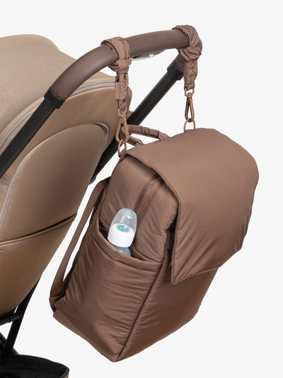 CALPAK Diaper Backpack with Laptop Sleeve attached to stroller by CALPAK Stroller Straps in hazelnut; BPB2401-HAZELNUT, BBPB2401-HAZELNUT