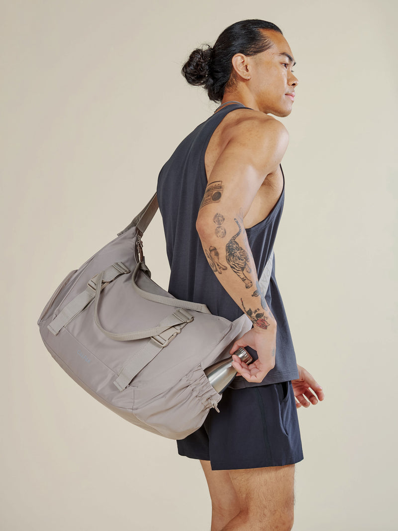 Model wearing grayish beige CALPAK Fit Gym bag as crossbody bag displaying side water bottle pocket with water bottle secured inside