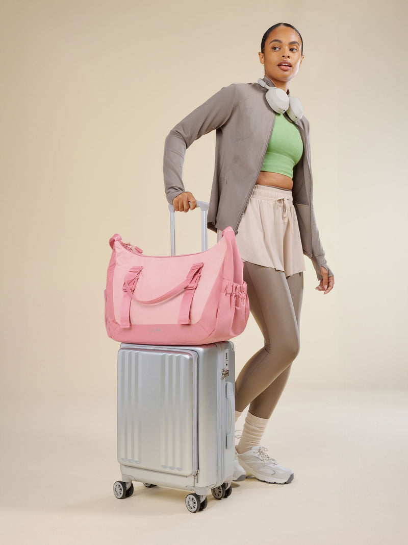 Model rolling CALPAK Ambeur Front Pocket Carry-On Luggage with tea rose pink CALPAK Fit Gym Bag secured by luggage trolley sleeve