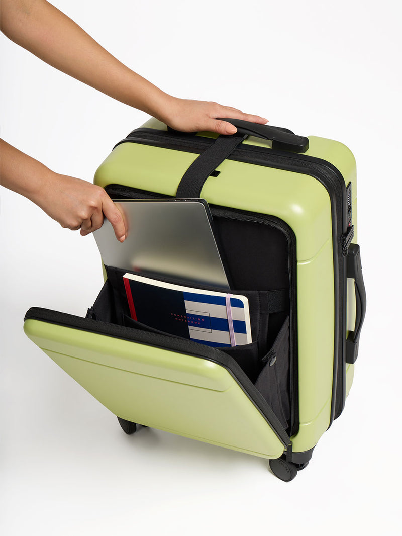 Model placing laptop and notebook in front pocket of CALPAK Hue Front Pocket Carry-On Luggage in green