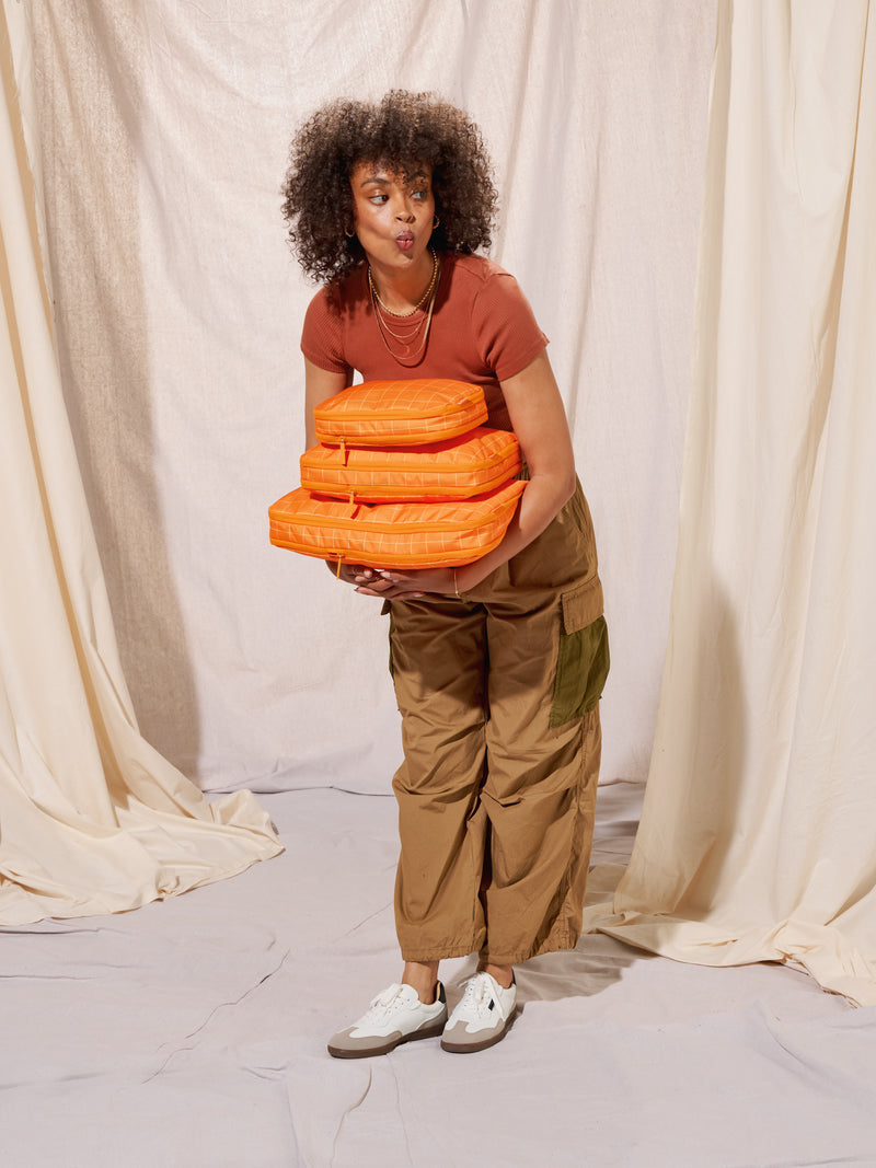 Model holding CALPAK Large Compression Packing Cube, Medium Compression Packing Cube, and Small Compression Packing Cube in orange grid