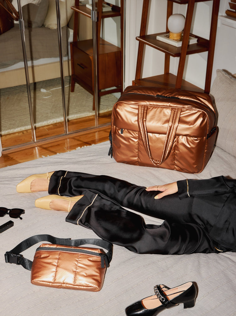 Model laying besides CALPAK Luka Duffel and CALPAK Belt Bag in metallic copper