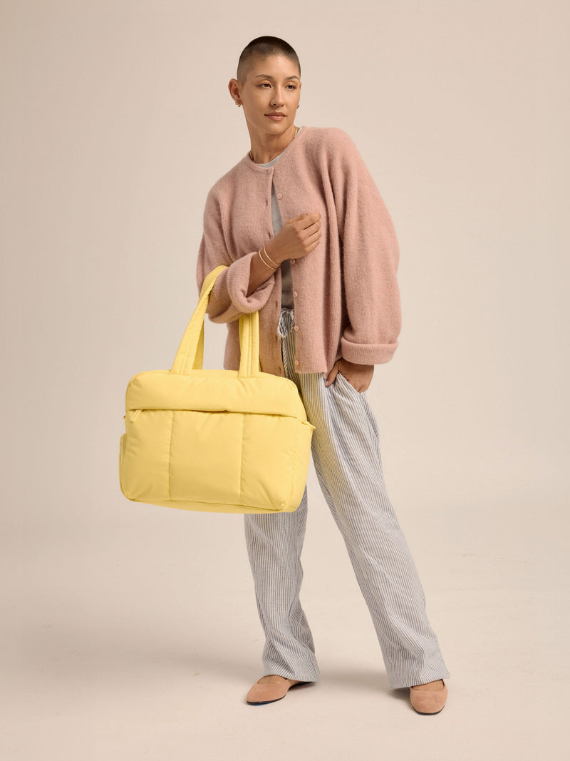 Model standing with yellow CALPAK Luka Duffel handle strap over arm