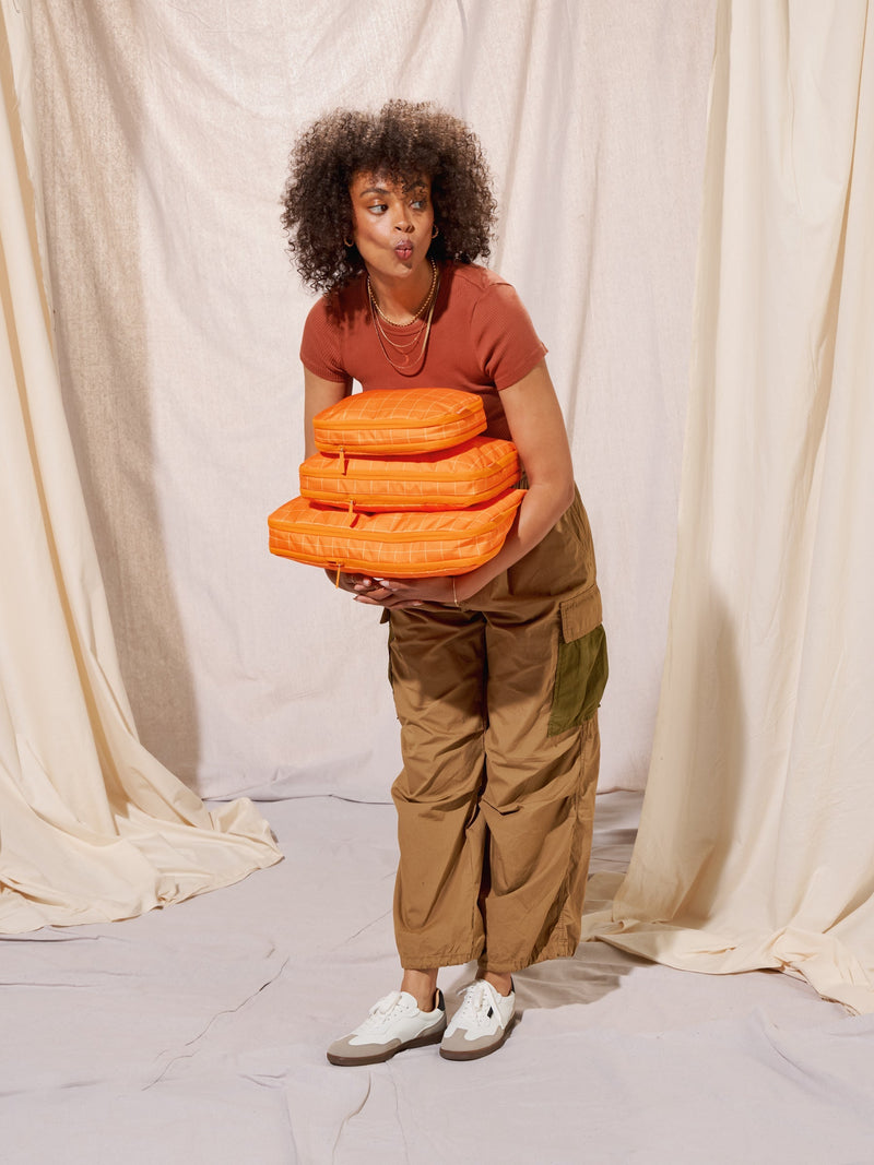 Model holding CALPAK Small Compression Packing Cube, CALPAK Medium Compression Packing Cube, and Large Compression Packing Cube in orange grid for size comparison