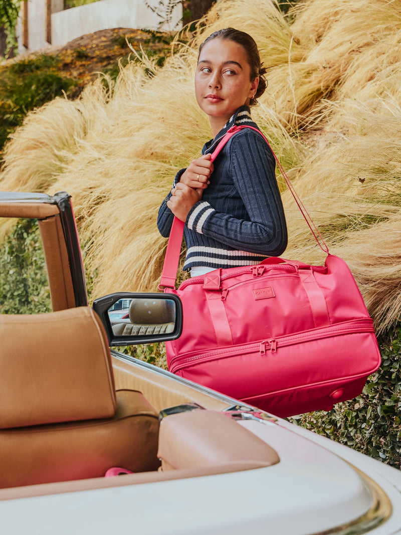 Model standing besides car holding dragonfruit CALPAK Stevyn Duffel with removable strap over shoulder