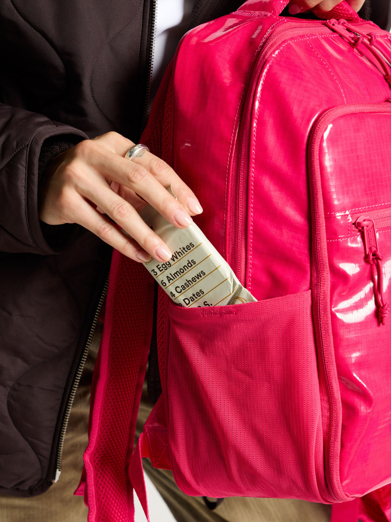 Model placing energy bar within the side pocket of CALPAK Terra Hydration Backpack in dragonfruit