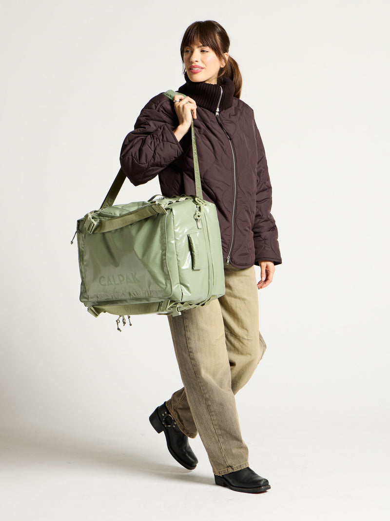 Model using adjustable shoulder strap to hold green CALPAK terra large 50L duffel backpack as duffel bag