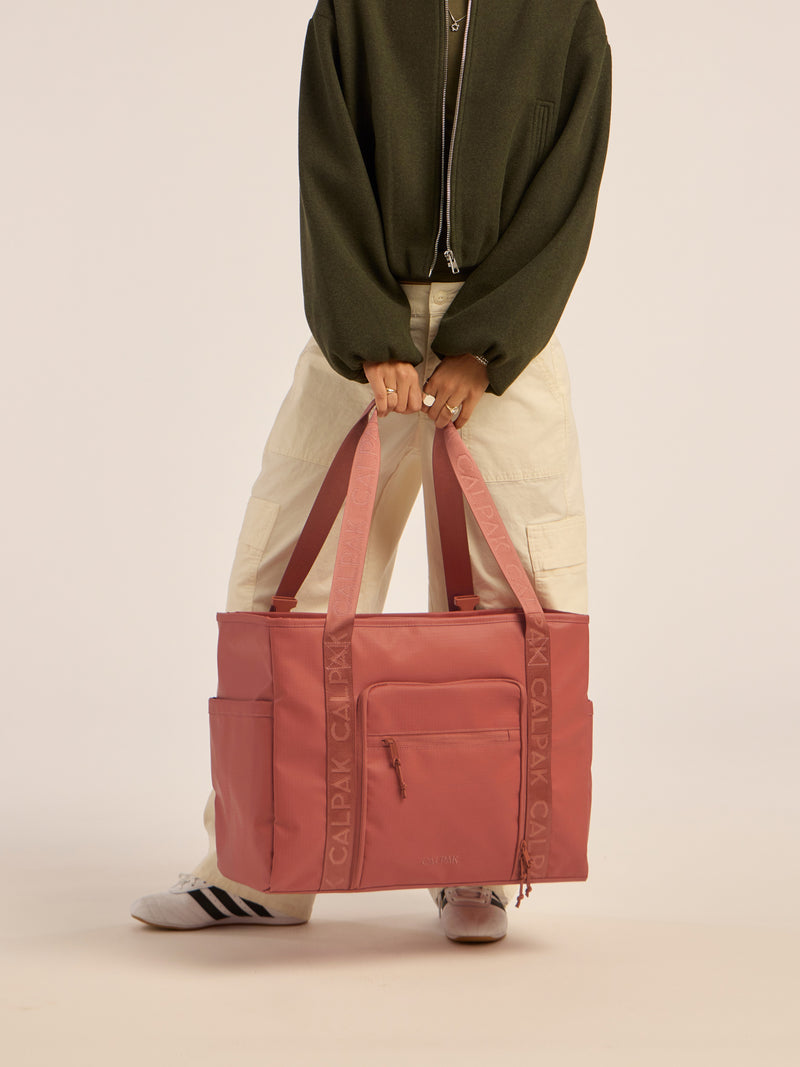 Model holding the CALPAK Terra 35L Water Resistant Zippered Tote Bag in red and orange Matte Clay