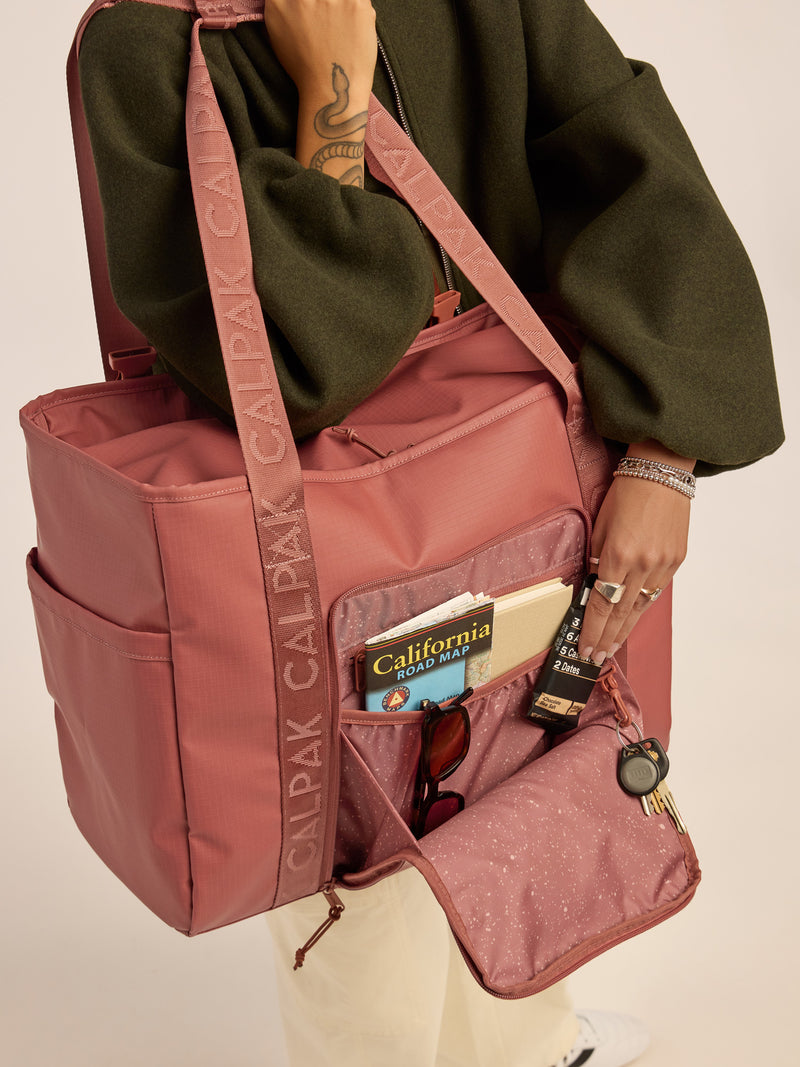 Model opening the front pocket of the CALPAK Terra 35L Water Resistant Zippered Tote Bag in red and orange Matte Clay