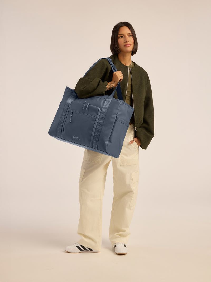 Model wearing CALPAK Terra 35L Water Resistant Zippered Tote Bag in blue Matte Indigo front view